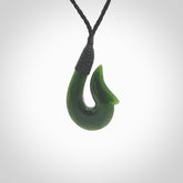 This photo shows a greenstone hook, or matau, pendant. It is a beautiful green British Columbia jade stone. The plaited cord is a black colour. It is plaited and the length can be adjusted.
